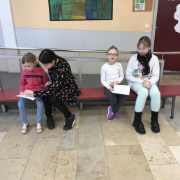 Zwei Erstklässlerinnen lesen – ältere Schülerinnen hören aufmerksam zu – Pausenhalle