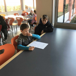 Junger Schüler liest aus der Fibel – älterer Schüler hört zu – Lesesituation in der Mensa