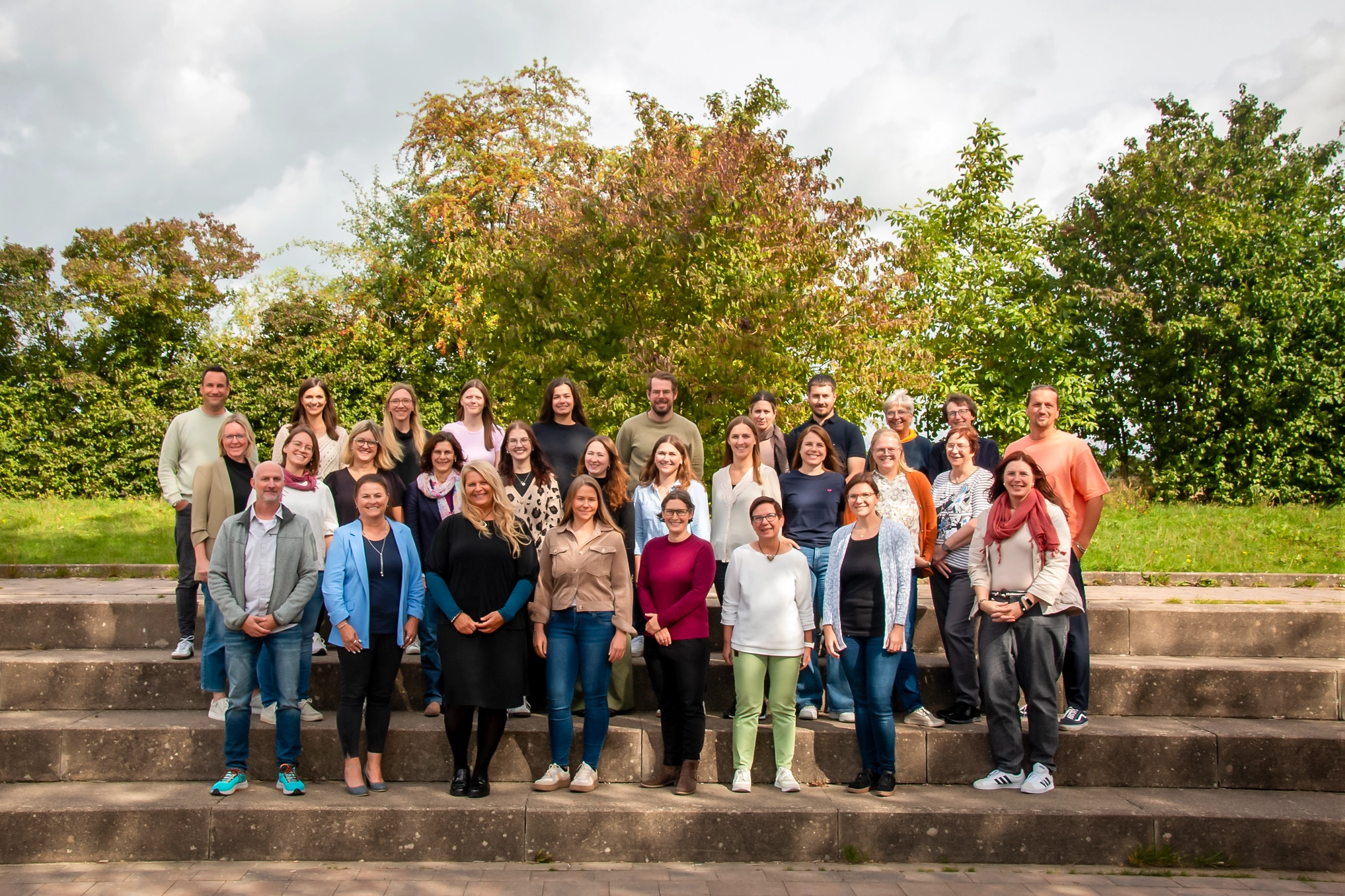 Gruppenfoto des Kollegiums im Freien, aufgestellte Lehrkräfte auf Steinstufen vor Bäumen.