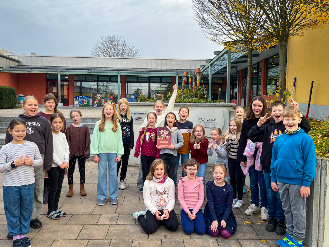 Eine große Gruppe fröhlicher Kinder steht auf dem Schulhof der Grund- und Mittelschule Bad Rodach. Sie lachen, jubeln und halten die Buchvorlage zum Musical „Der Beat deines Lebens“ hoch. Im Hintergrund sind Schulgebäude, herbstliche Bäume und bemalte Elemente des Schulgeländes zu sehen.