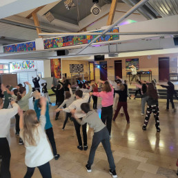 Schüler & Lehrer tanzen in der Aula und folgen den Bewegungen der Tanzlehrerin auf der Bühne.
