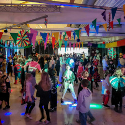 Viele verkleidete Kinder tanzen in einer mit Girlanden dekorierten Schulhalle unter bunten Lichteffekten.