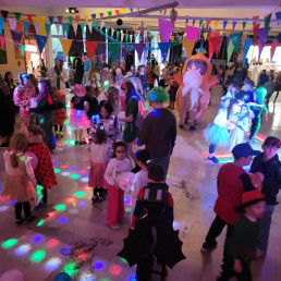 Verkleidete Kinder stehen und tanzen mit Luftballons in einer festlich beleuchteten Schulhalle.