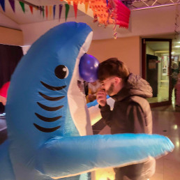 Eine Person im aufblasbaren Hai-Kostüm steht während der Faschingsfeier zusammen mit einem Schüler in der geschmückten Schulhalle.